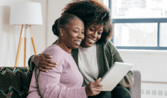 A black woman puts her arm around her elder mother as they look at an ipad