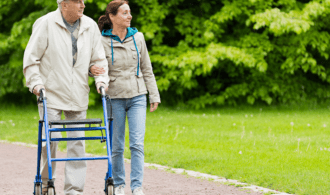 An older white man in a hat walks next to his daughter using a walker.