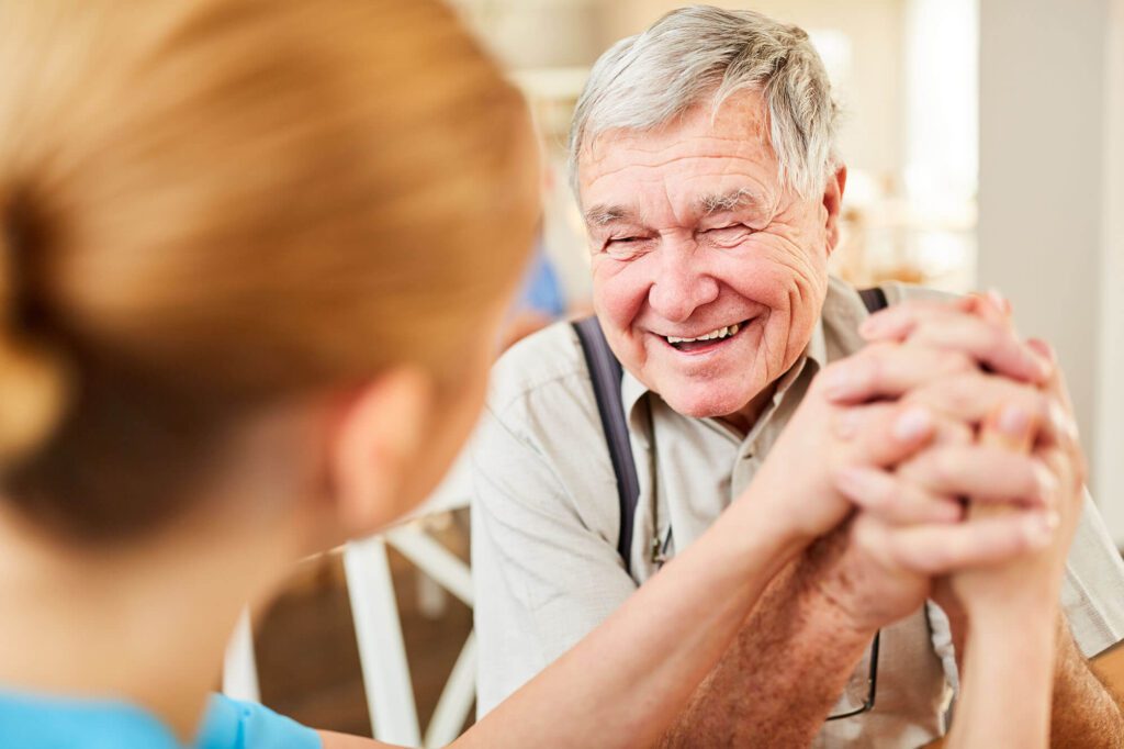 a elderly man holds the hands of a caretaker with a big smile
