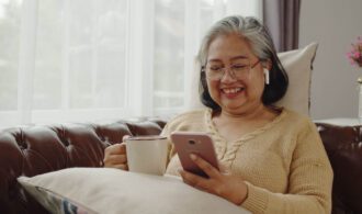 A woman in assisted living sits on a couch with her cell phone and a cup of tea
