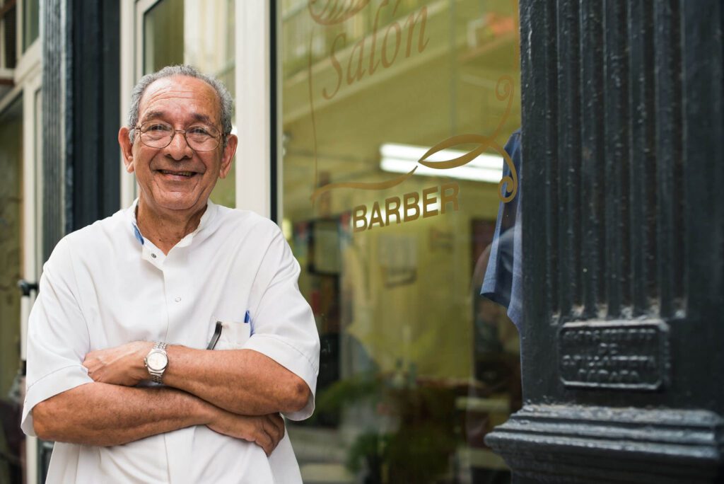 an elderly man stands in front of a barber business with a big grin