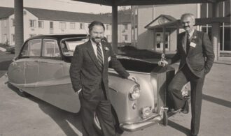 black and white photograph of 2 men stand in front of a car from the 50s
