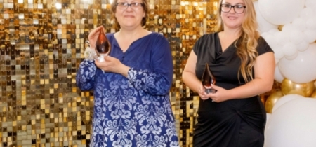 Two women accept an award in front of a gold backdrop